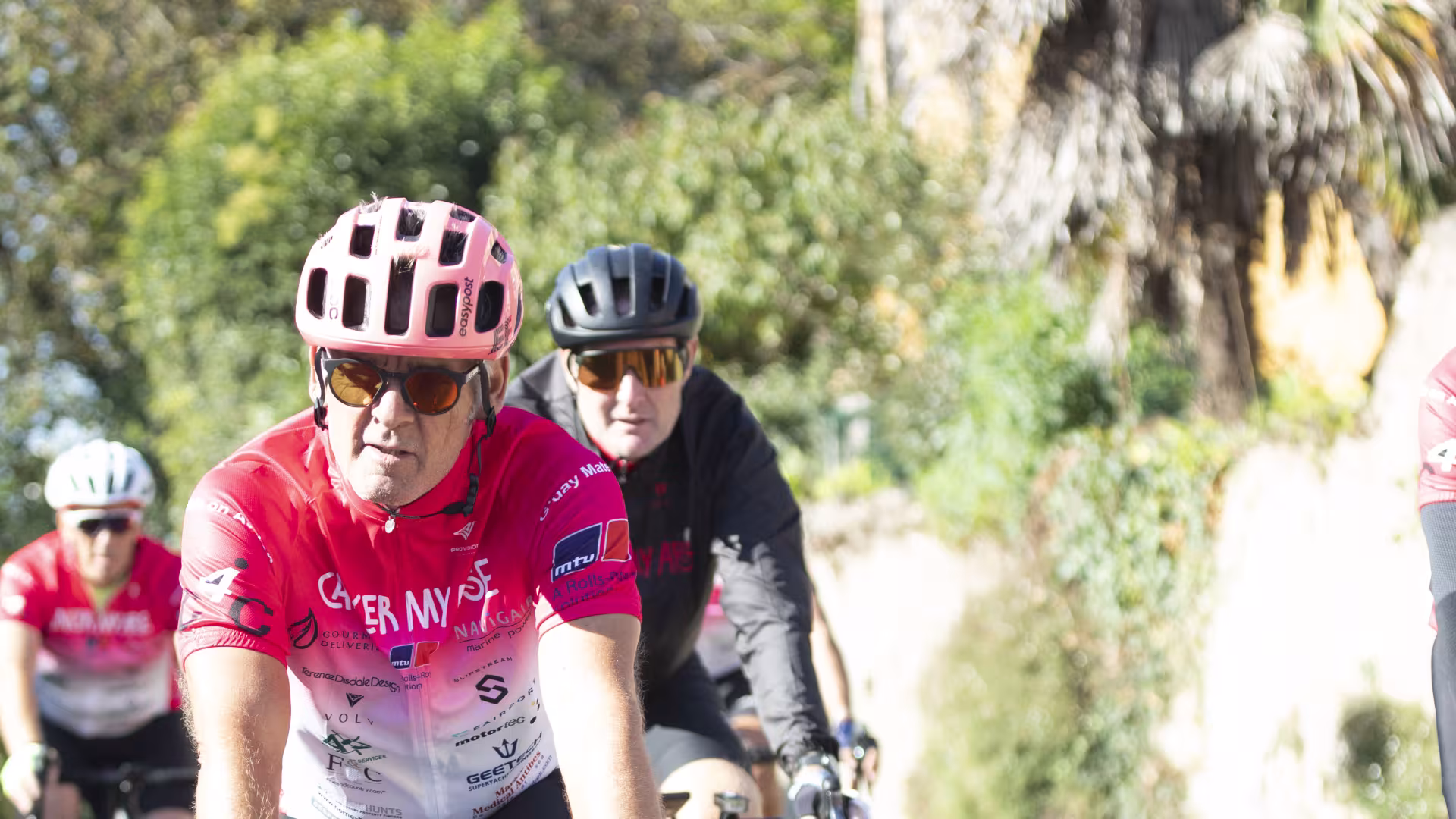 Three riders in pink Cancer My Arse kit cycling along a sunny tree-lined road