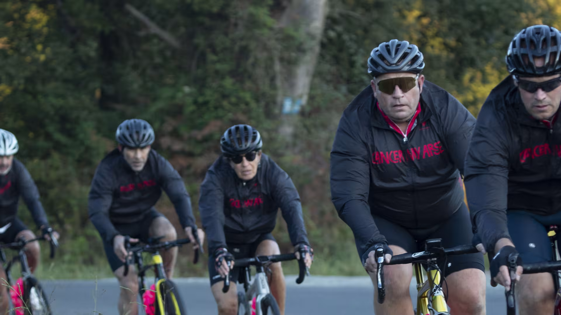 Five riders in black and pink Cancer My Arse jackets cycling along a misty tree-lined road